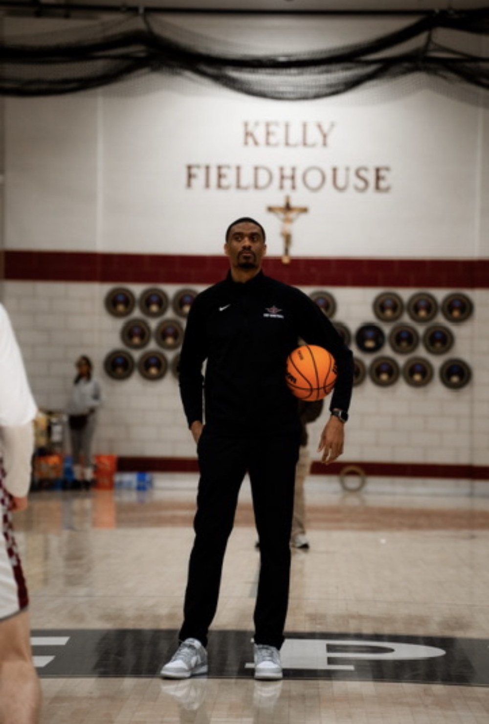 Earl Brown Jr. standing at center court in a gym, wearing a black quarter-zip and holding a basketball, with championship plaques mounted on the wall behind him.